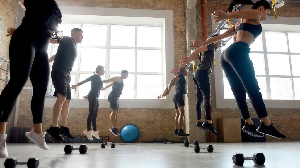 Group of sportive people jumping, warming up before having workout with dumbbells at industrial gym. Crossfit group training concept. Horizontal shot. Side view